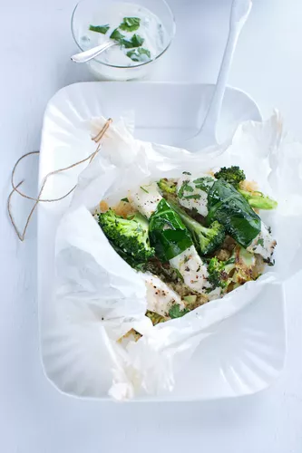Fotoproduktion Auf weißem Geschirr präsentiert, in Papier eingewickelter, gekochter Fisch mit Kräutern und Gemüse. Dazu ein Schälchen mit weißer Soße.
